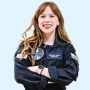 A smiling woman with long reddish brown hair and her arms folded over her space jumpsuit