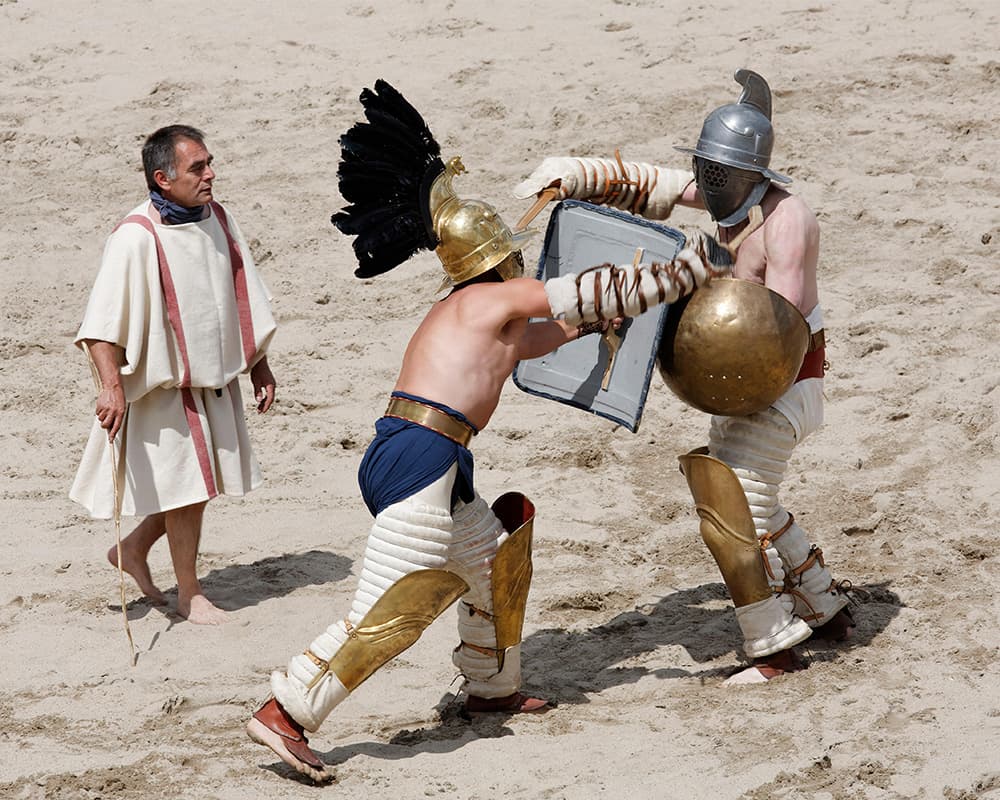 Two men with metal warrior helmets battling each other with swords in the sandy arena